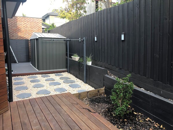 A wooden deck with stepping stones leading to a shed, surrounded by a black fence and a brick wall.