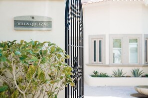 Uma entrada com portão, placa indicando 'VILLA QUETZAL' e um vaso de planta em primeiro plano.