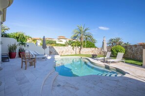 Piscina com espreguiçadeiras e guarda-sóis, cercada por um muro de pedra e palmeiras.