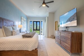 A bedroom with a large bed, a wooden dresser, a ceiling fan, and a TV on the wall.