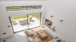 Ett modernt vardagsrum med en grå soffa, ett soffbord och en tv som är monterad på väggen. Det finns en skjutdörr i glas som leder ut till en uteplats med möbler i rotting.