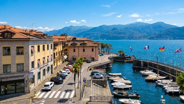 Una calle frente al mar con coches aparcados, un puerto deportivo con barcos y edificios con balcones.