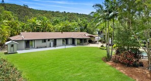 Une maison moderne à deux étages, dotée d’un toit en tuiles, entourée d’une pelouse bien entretenue et d’une végétation tropicale.