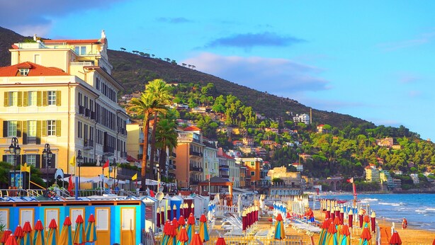 Uma vila costeira com edifícios coloridos, uma praia com guarda-sóis e uma colina ao fundo.