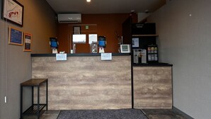 A reception area with a wooden reception desk, two monitors, a small table, and a grey carpet. - HOTEL R9 The Yard Iwakuni (Iwakuni)