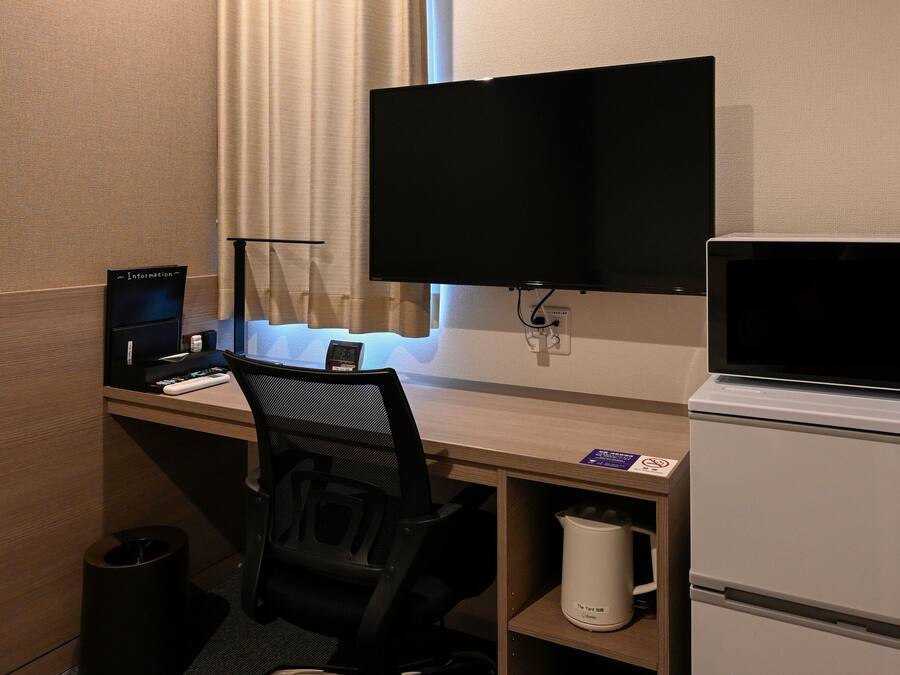 A hotel room with a desk, a computer, a TV, a microwave, and a trash can.