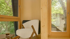 Una habitación acogedora con un ventanal grande, suelo de madera, una silla blanca y una mesita con un vaso.