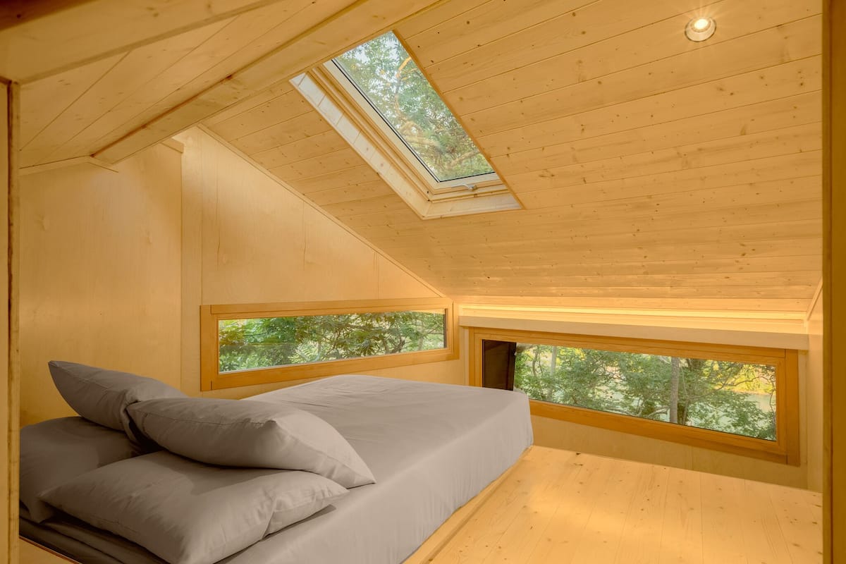 Un dormitorio con tragaluces, una cama con almohadas y ventanas con vistas a árboles.