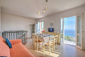 Ampla sala de estar com uma mesa de jantar para quatro pessoas, sofá, varanda com vista e cozinha ao fundo.
