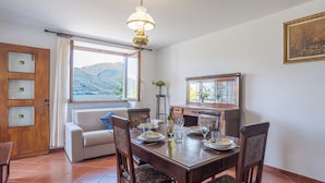 A dining area with a wooden table set for four, a sofa, and a view of mountains through a window. - Casa Achille Pianello (Pianello del Lario)