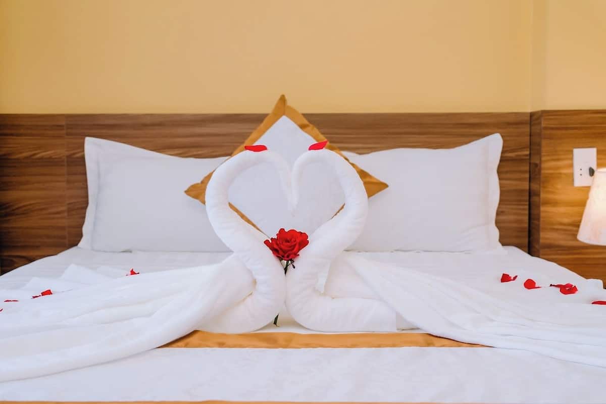 A bed with a white bedspread and pillows, a wooden headboard, and a red rose placed in the center.