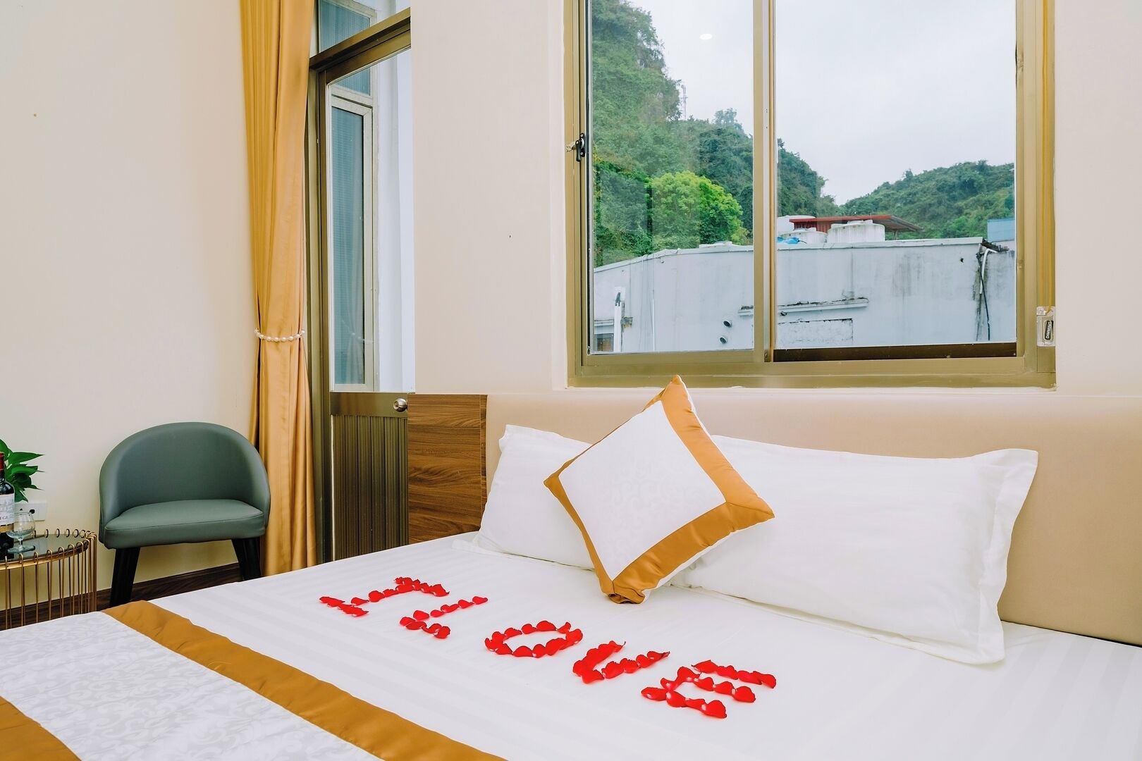 A hotel room with a bed, a chair, a window with a view of buildings and greenery, and a door.