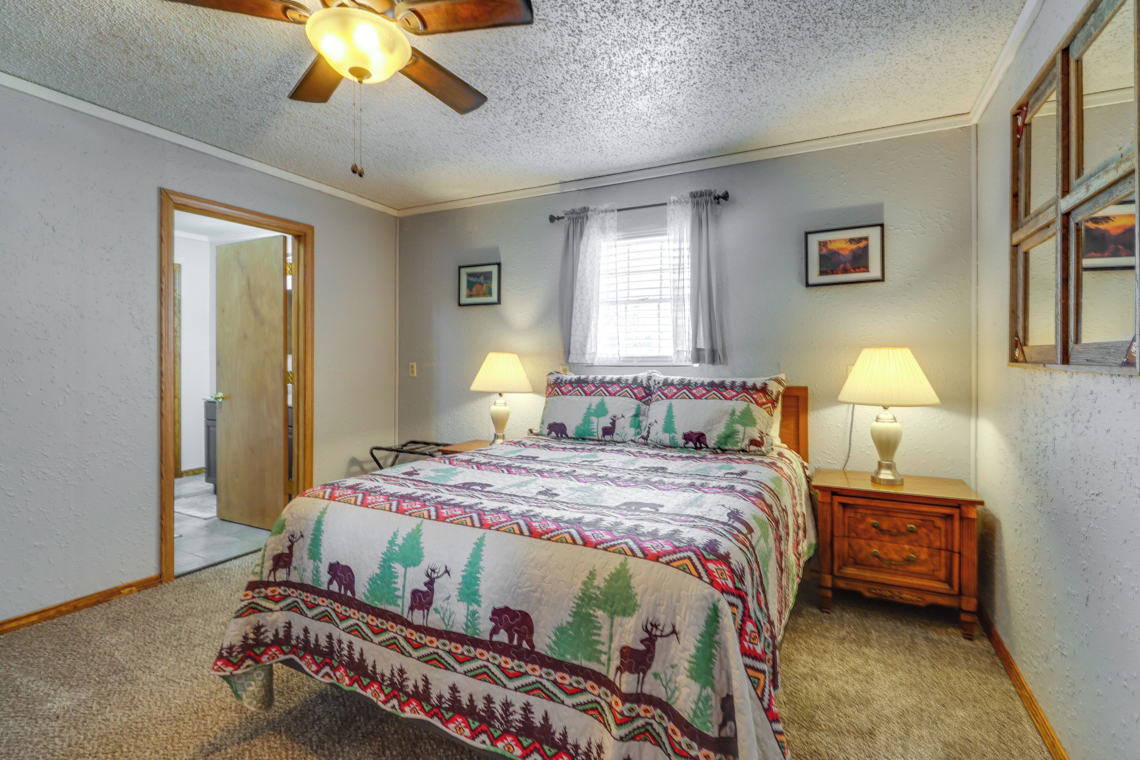 A bedroom with a bed, two bedside tables with lamps, a ceiling fan, a window with curtains, and a door leading to another room.
