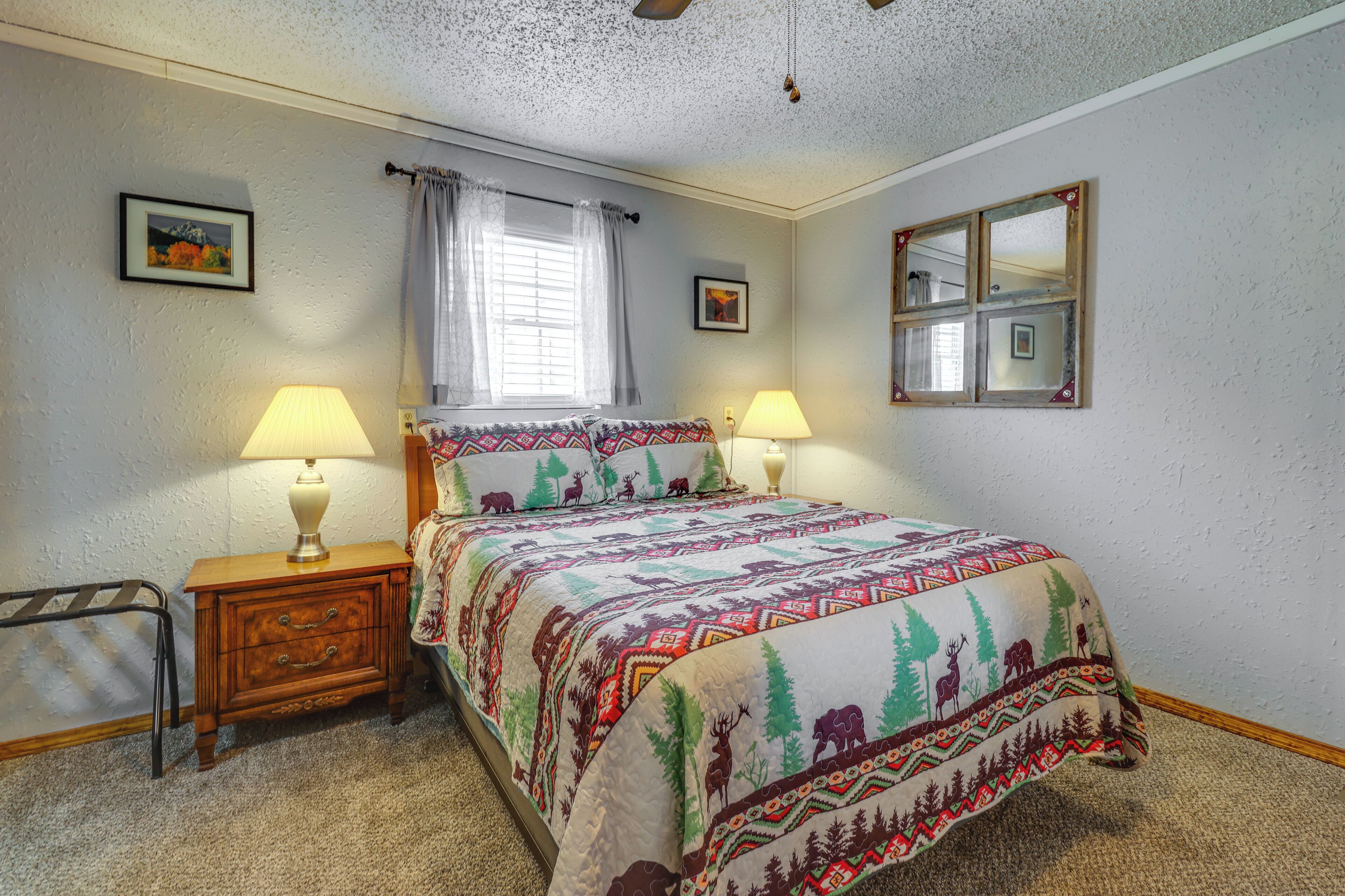 A bedroom with a bed, two bedside tables, a nightstand, a window with curtains, a wall-mounted mirror, and two framed pictures.