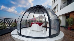A covered outdoor dining area with a table and chairs, surrounded by a circular glass enclosure. - Shanti seaview resort and spa (Kanthi)