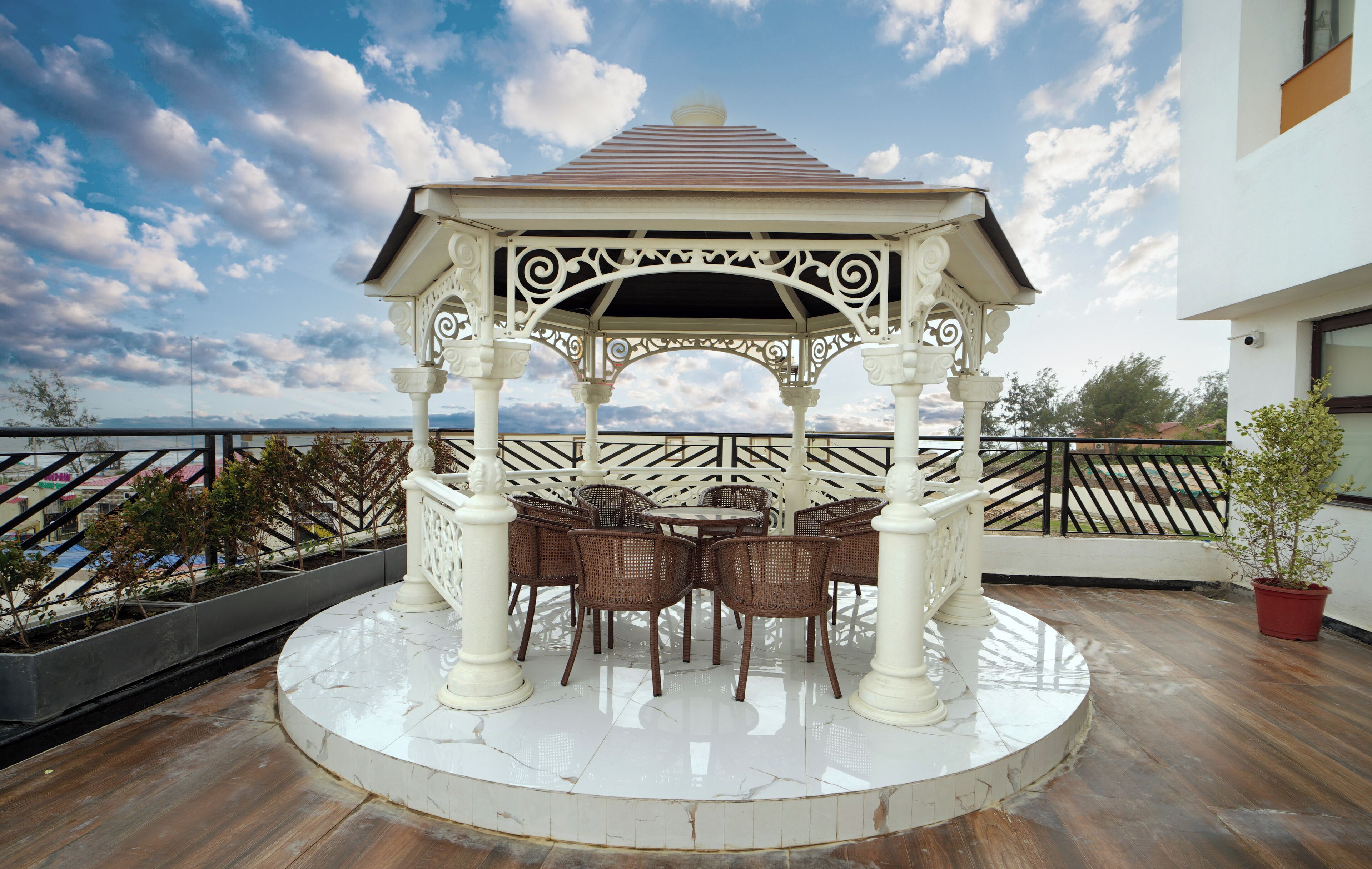 Un pabellón con techo blanco y diseño ornamentado, rodeado de sillones de mimbre y una mesa blanca, situado en un balcón con vistas al mar.