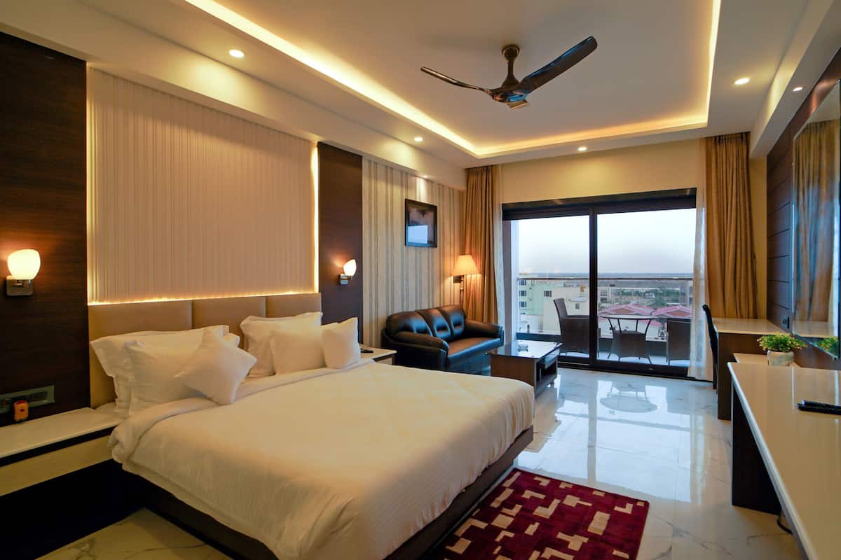 A modern hotel room with a large bed, a seating area with a sofa and armchairs, a dining table, and a balcony with a view.