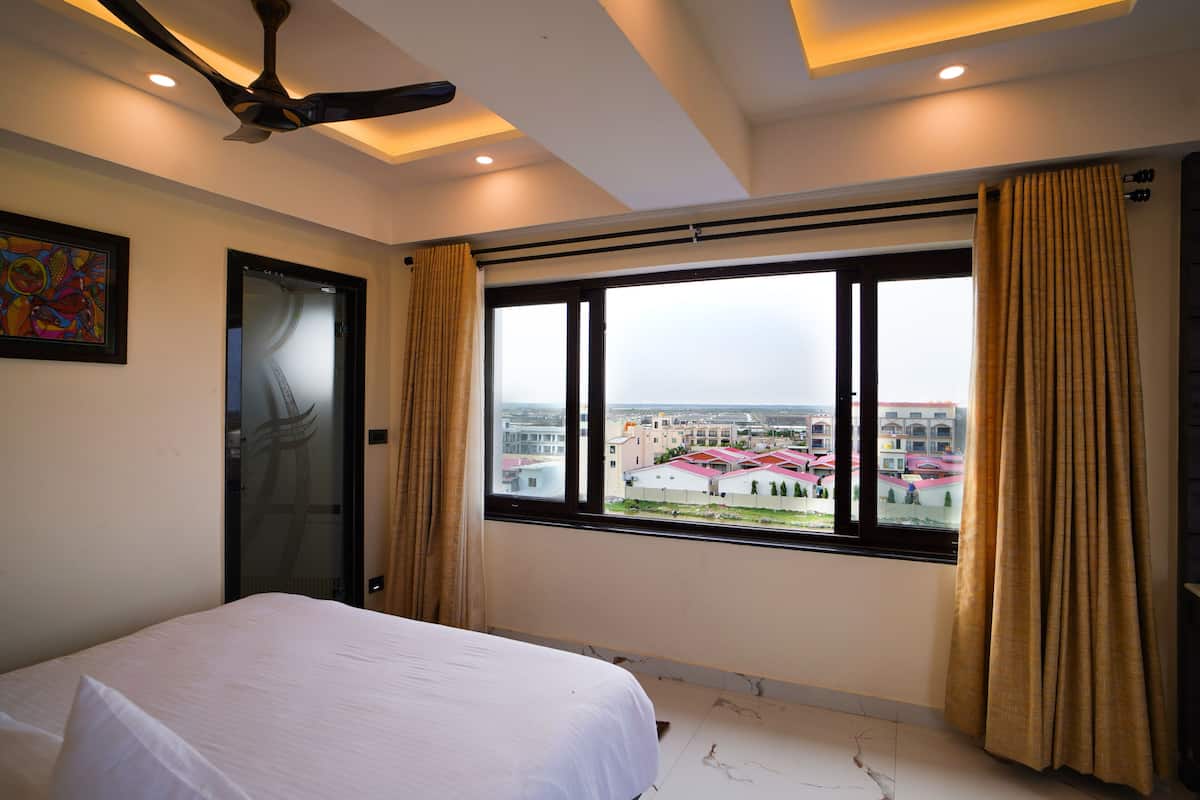 A hotel room with a bed, a ceiling fan, a window with curtains, and a view of buildings outside.