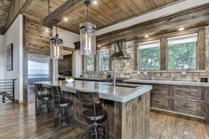 A kitchen with a large island, bar stools, a stone backsplash, and a view through the windows.
