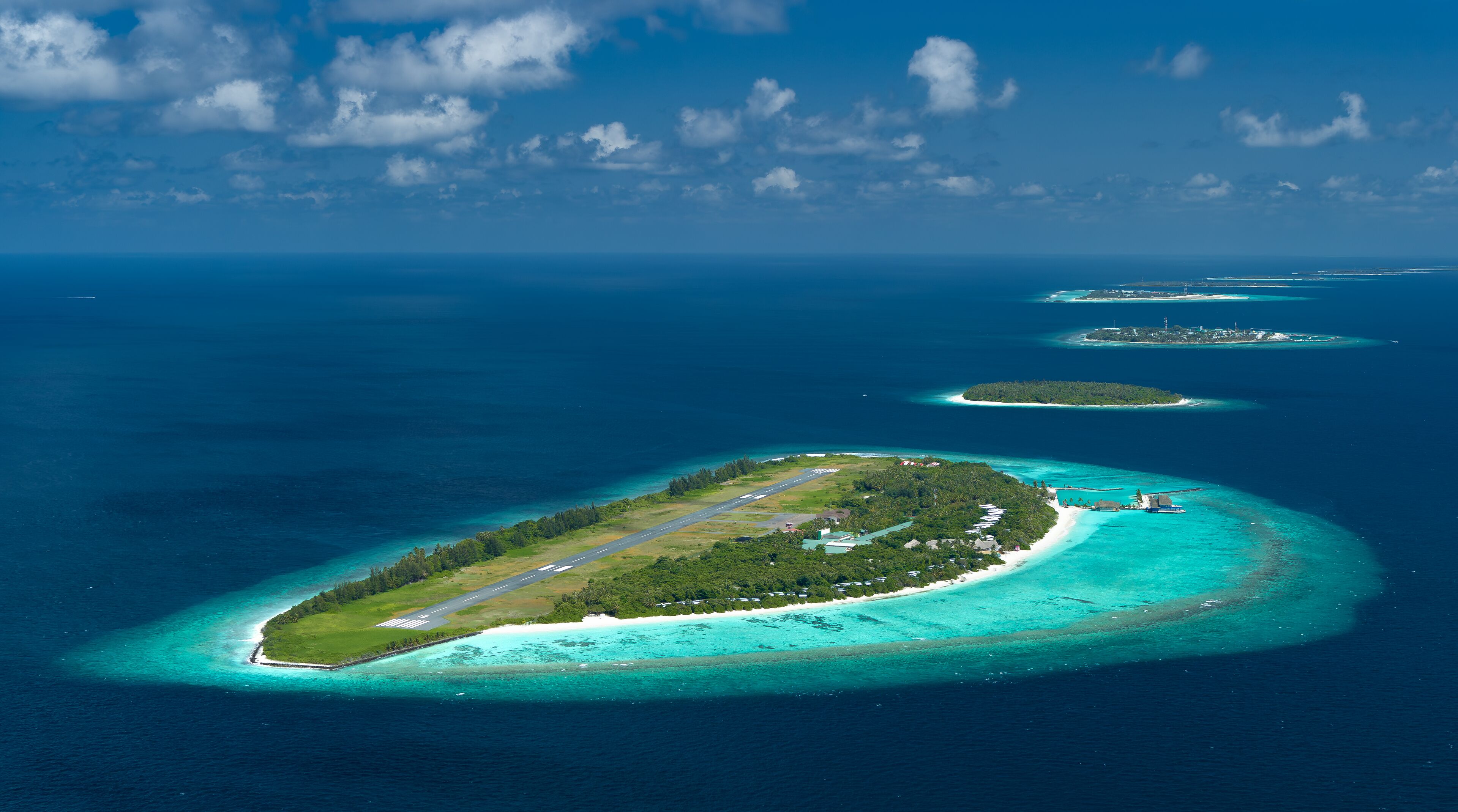 An aerial view of a tropical island with a runway, surrounded by turquoise waters and smaller islands in the distance.