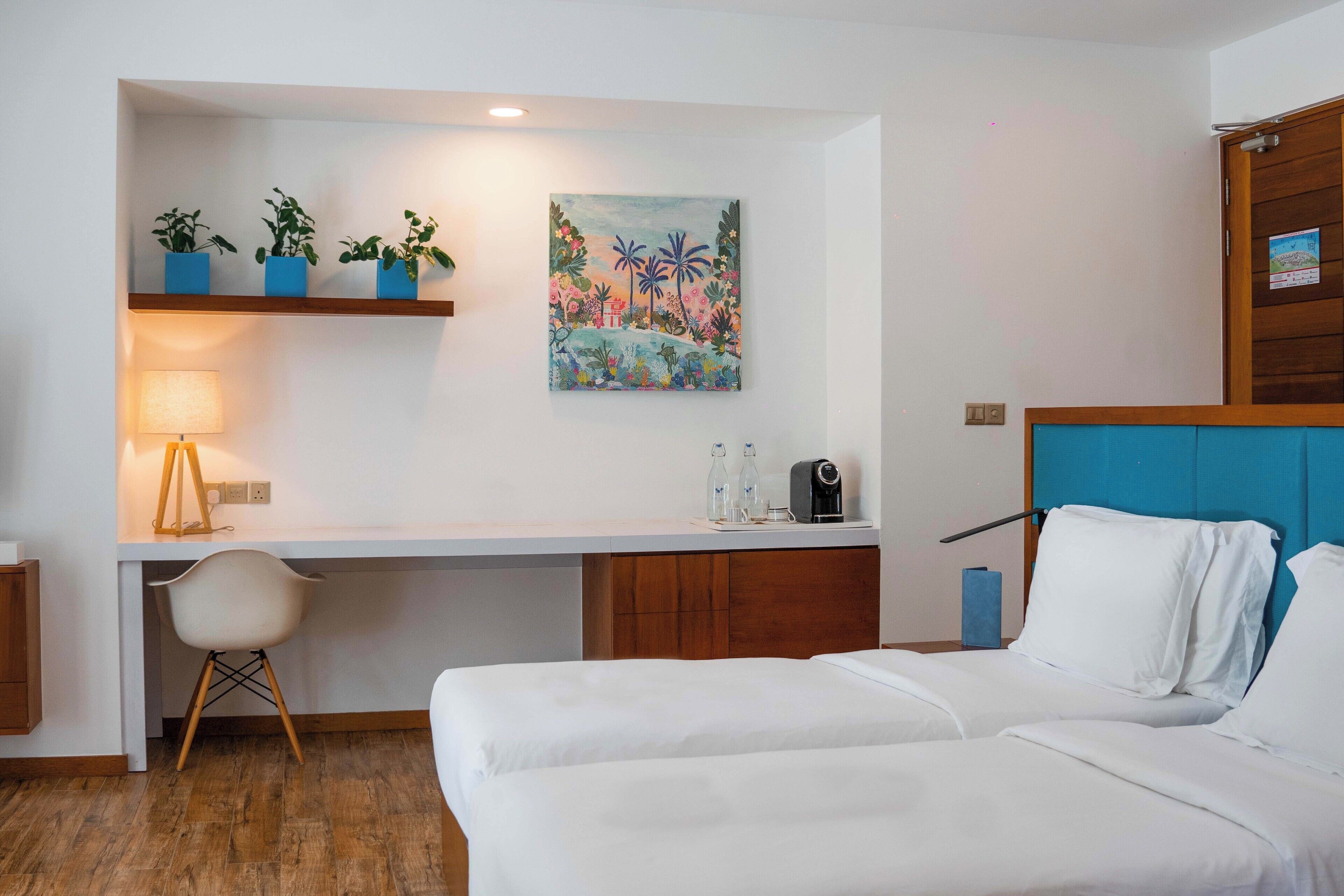 Habitación de hotel con una cama, un escritorio con silla, una lámpara, un cuadro y tres macetas con plantas sobre una estantería.