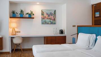 Habitación de hotel con una cama, un escritorio con silla, una lámpara, un cuadro y tres macetas con plantas sobre una estantería.