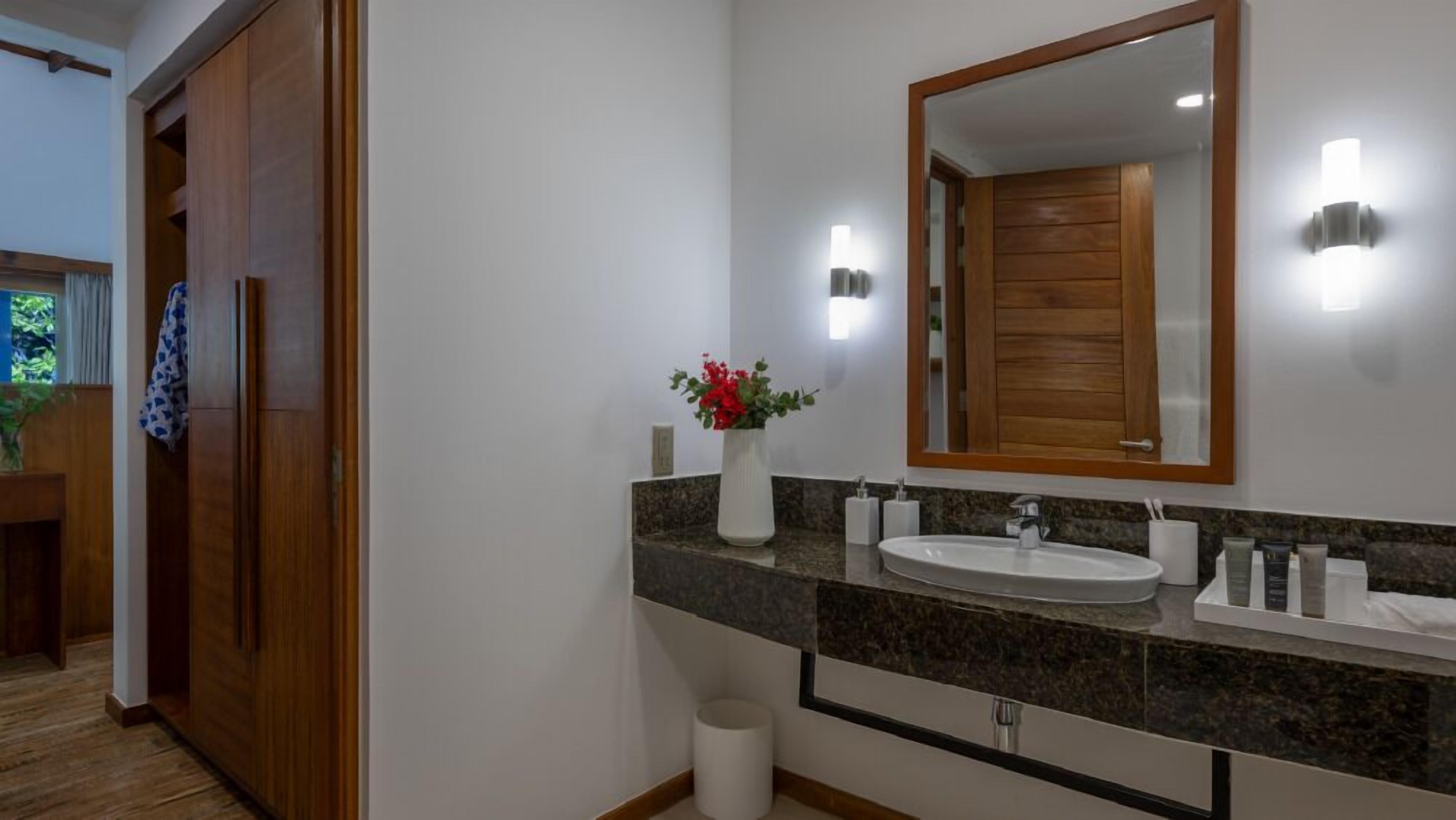 Ein Badezimmer mit Holztür, weißem Waschbecken, einem Spiegel und einer Vase mit roten Blumen.