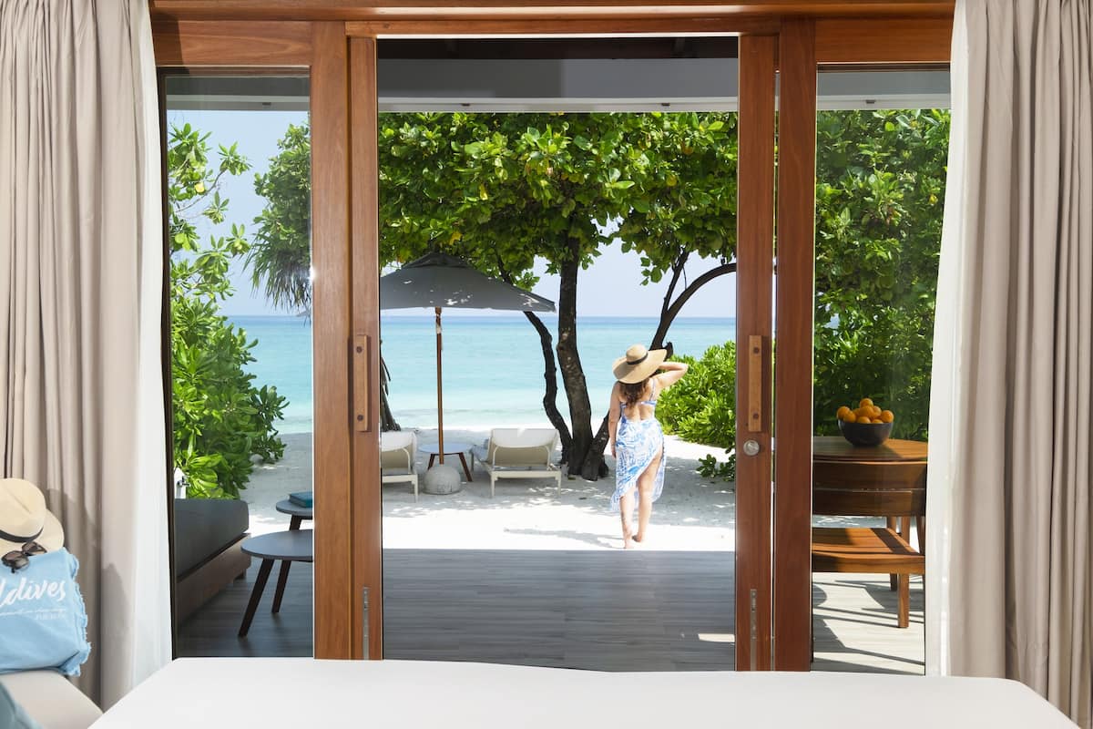 A bedroom with a view of a beach, a wooden deck, and outdoor seating.