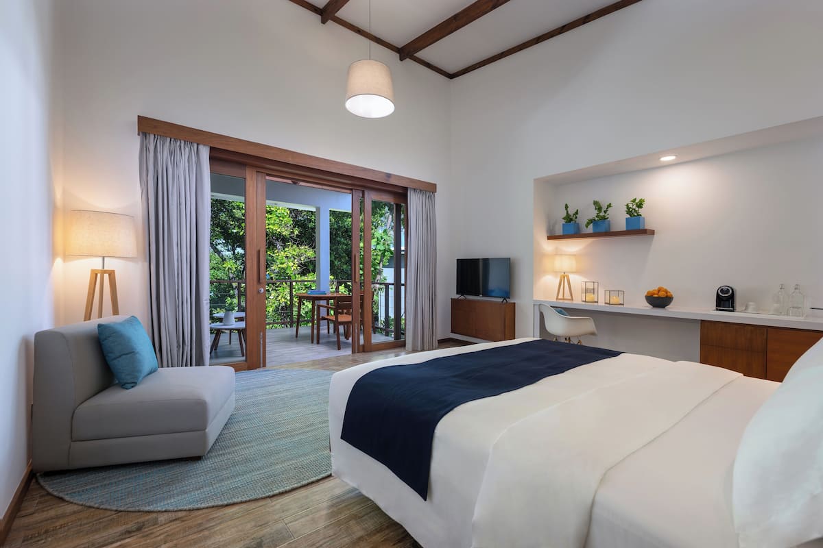 A modern hotel room with a large bed, a sofa, a desk, and a TV. There is a balcony with a table and chairs, and a view of greenery outside.