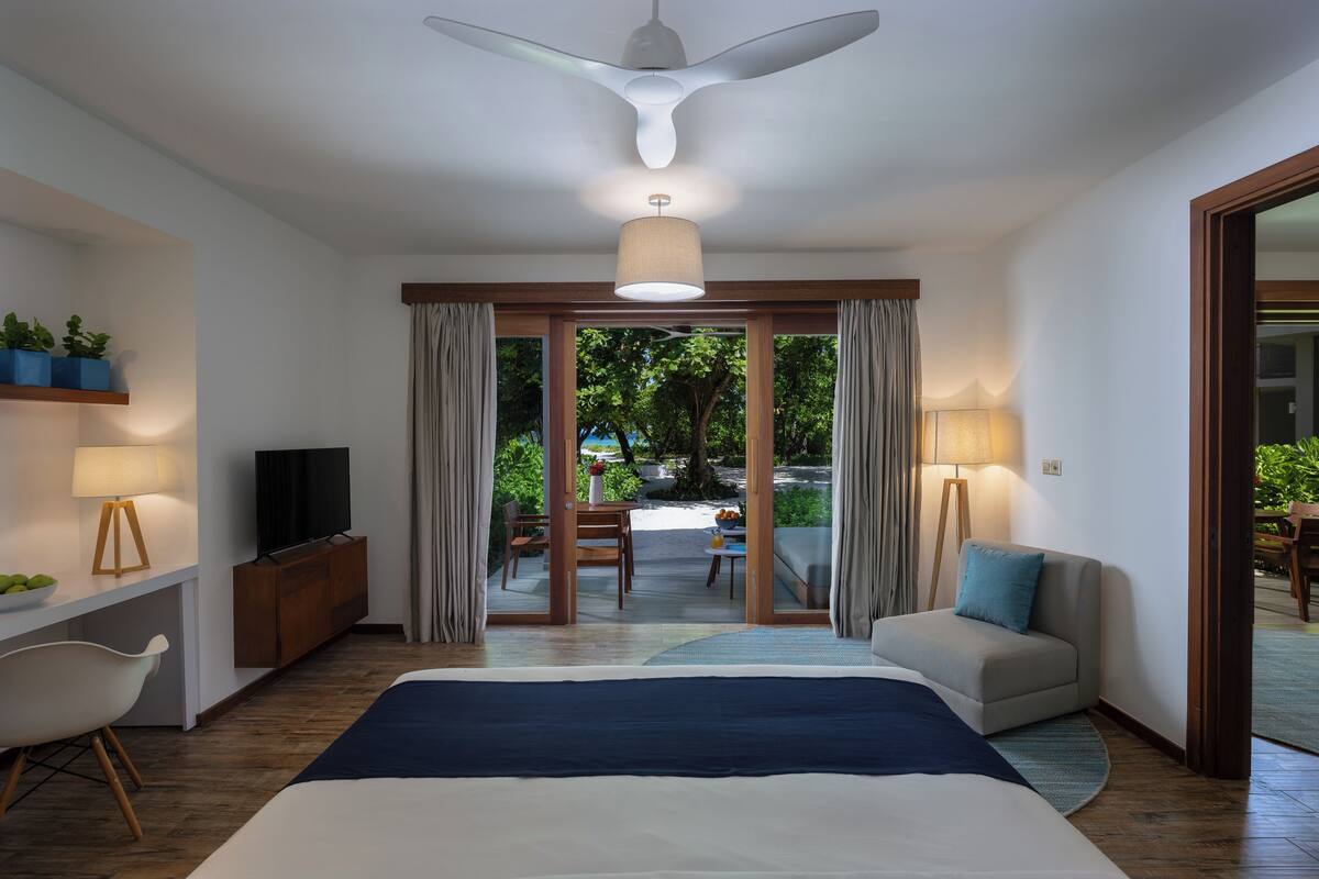 A modern hotel room with a large bed, a TV, a desk with a lamp, a chair, and a view of the outdoor area through a glass door.