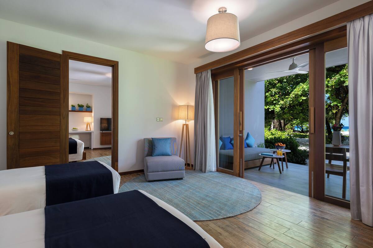 A hotel room with two beds, a sitting area with a lamp, and a view of an outdoor deck.