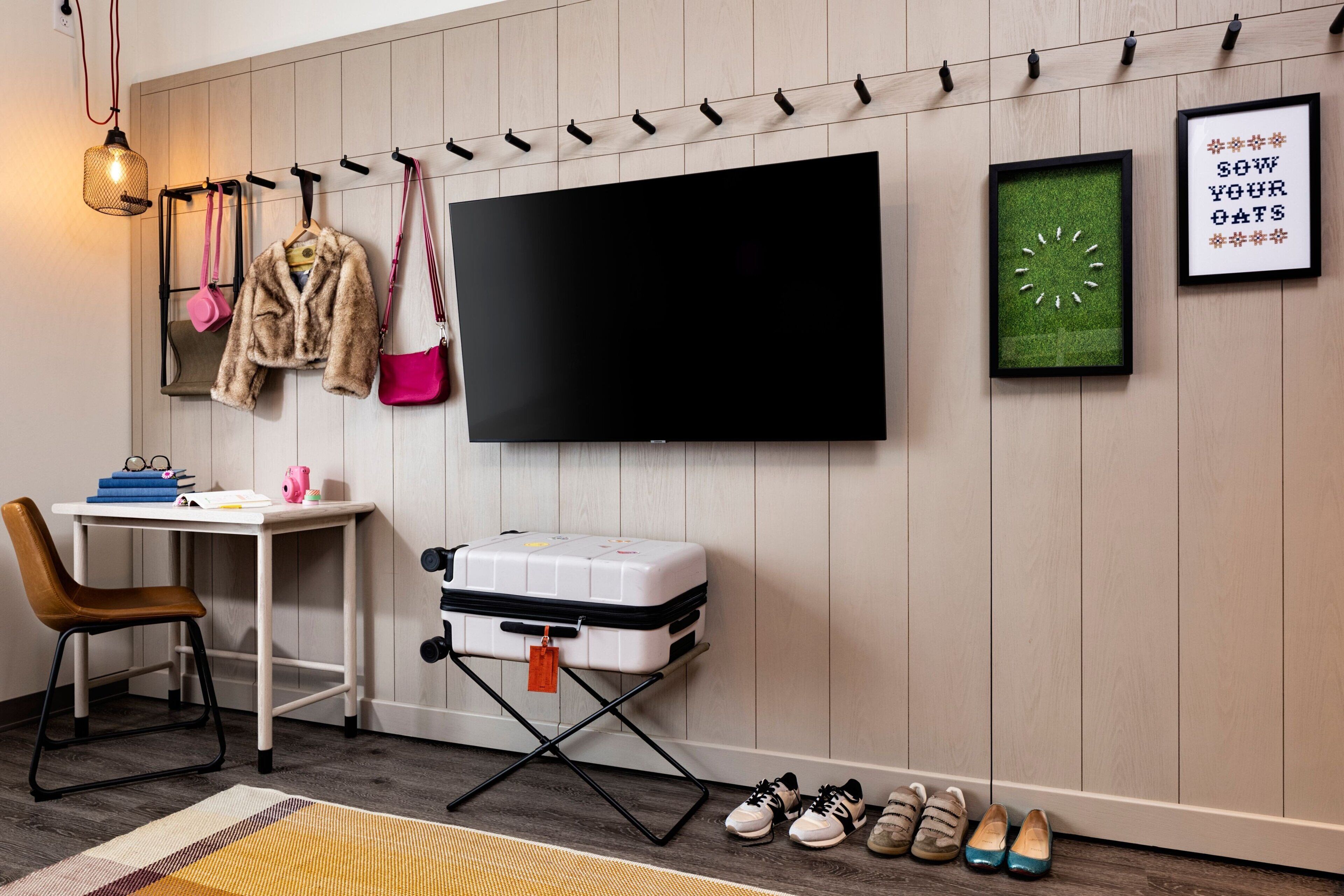 Ein Zimmer mit einem an der Wand montierten Flachbildfernseher, einem Schreibtisch mit Koffer, einem Stuhl und einem Paar Schuhe.