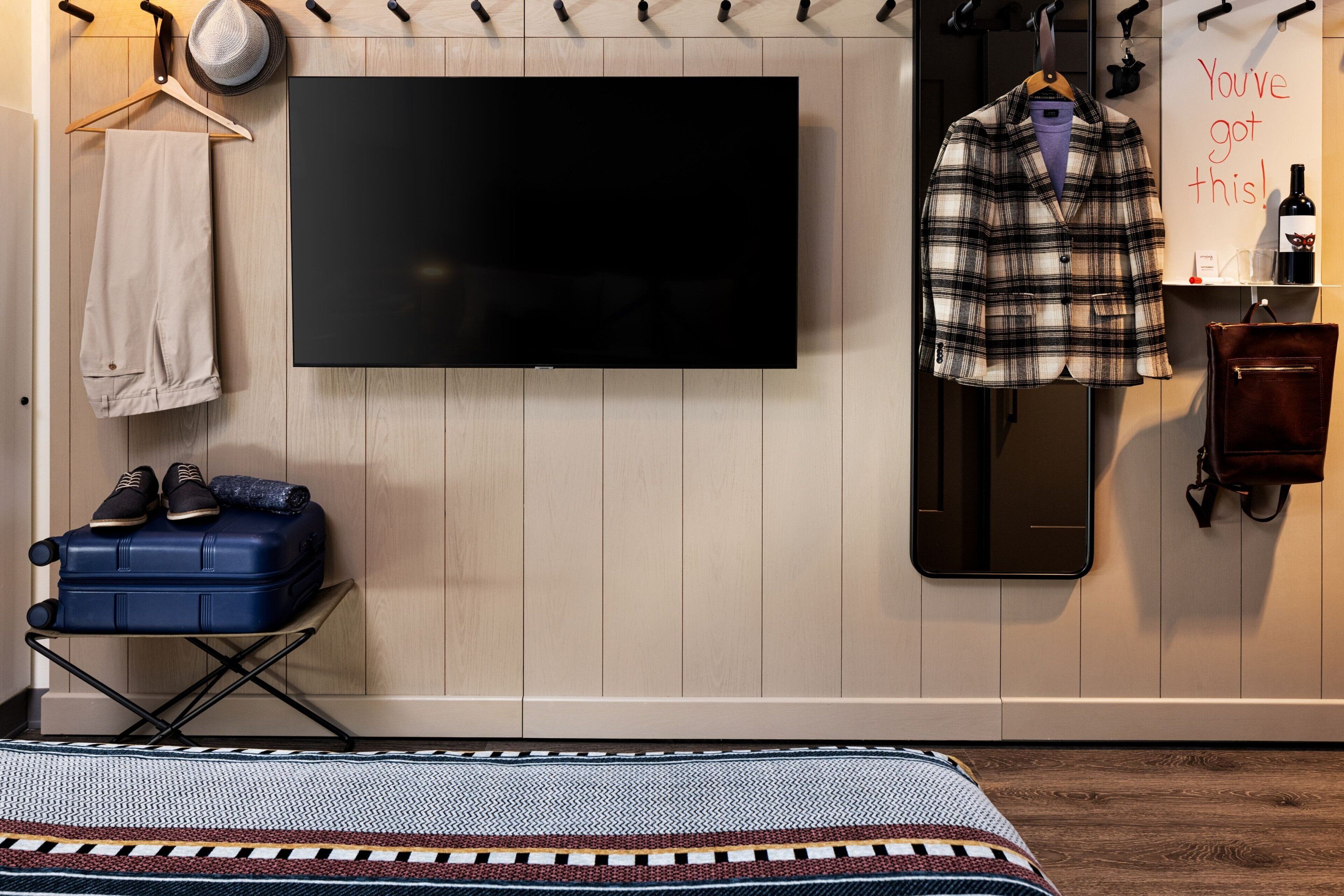 A room with a large flat-screen TV mounted on the wall, a suitcase on a small table, a plaid jacket hanging on a rack, and a sign that reads "You've got this!"