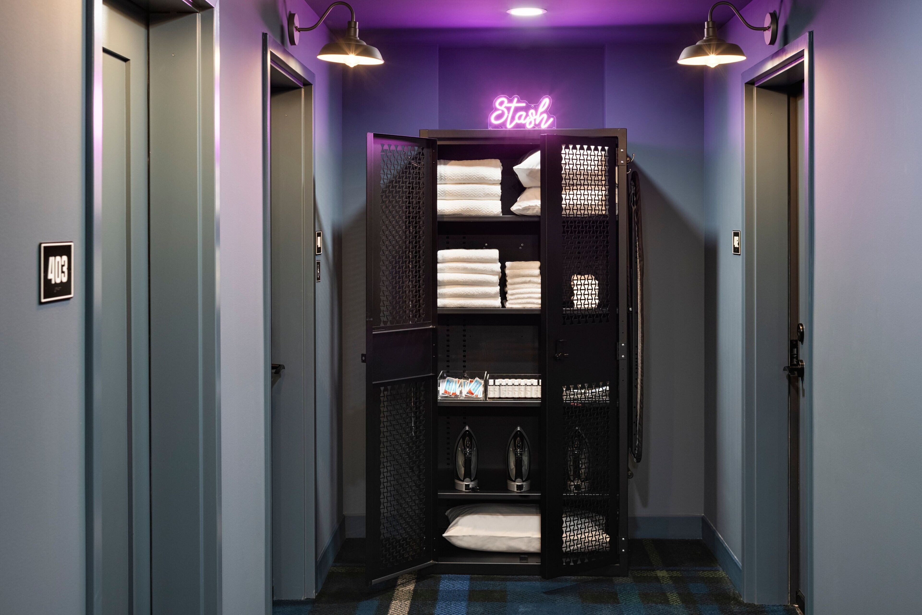 A hallway with an elevator, a neon sign reading "Stosh," and a storage unit with towels and irons.
