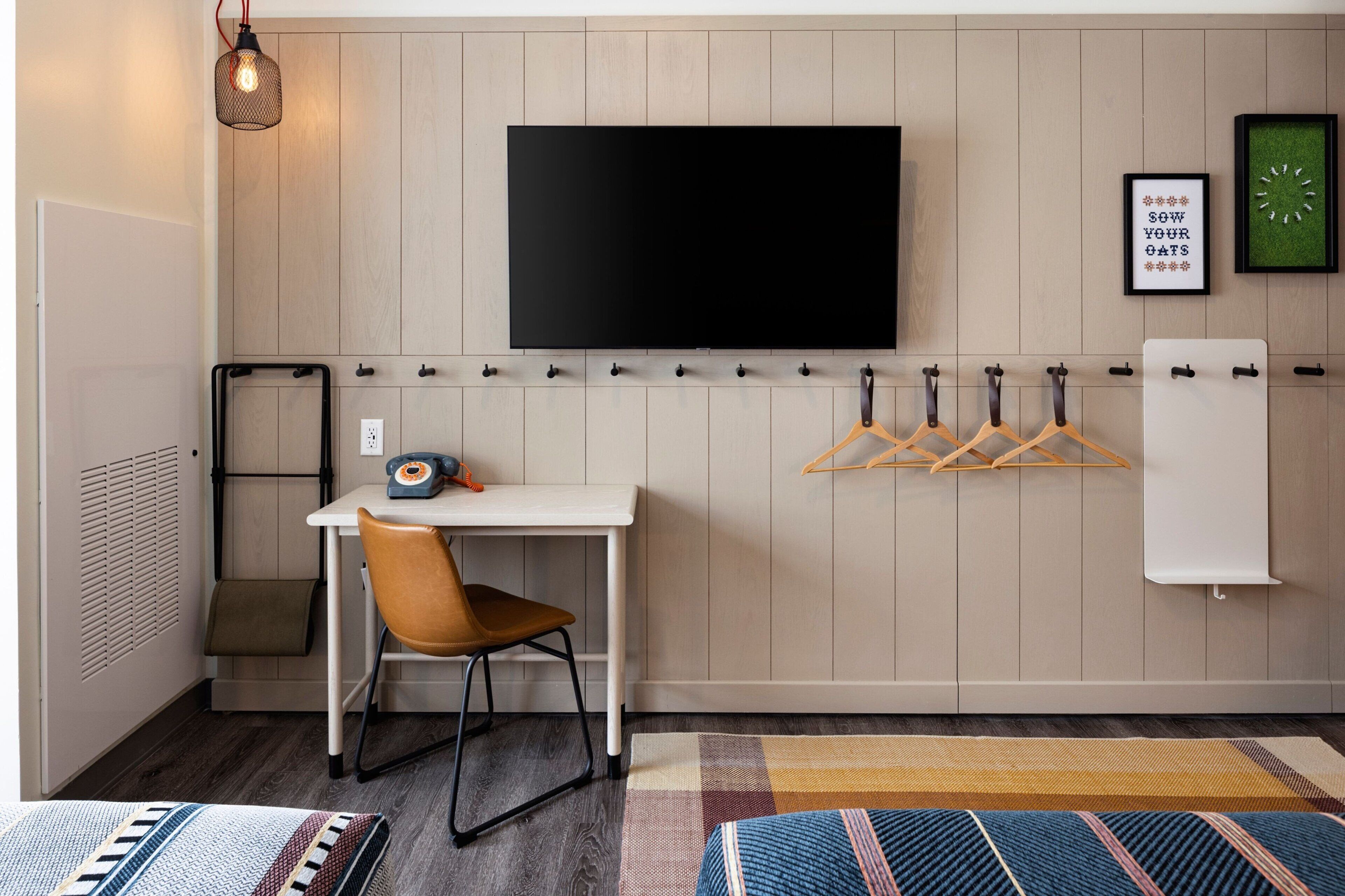 A modern room with a mounted TV, a desk with a chair, a coat rack, and a bed with a colorful rug.