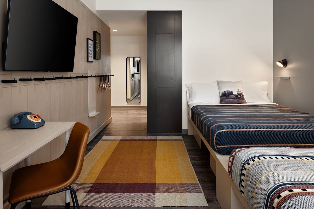 A modern hotel room with a flat-screen TV, a desk with a vintage telephone, a large bed with patterned bedding, and a colorful rug.