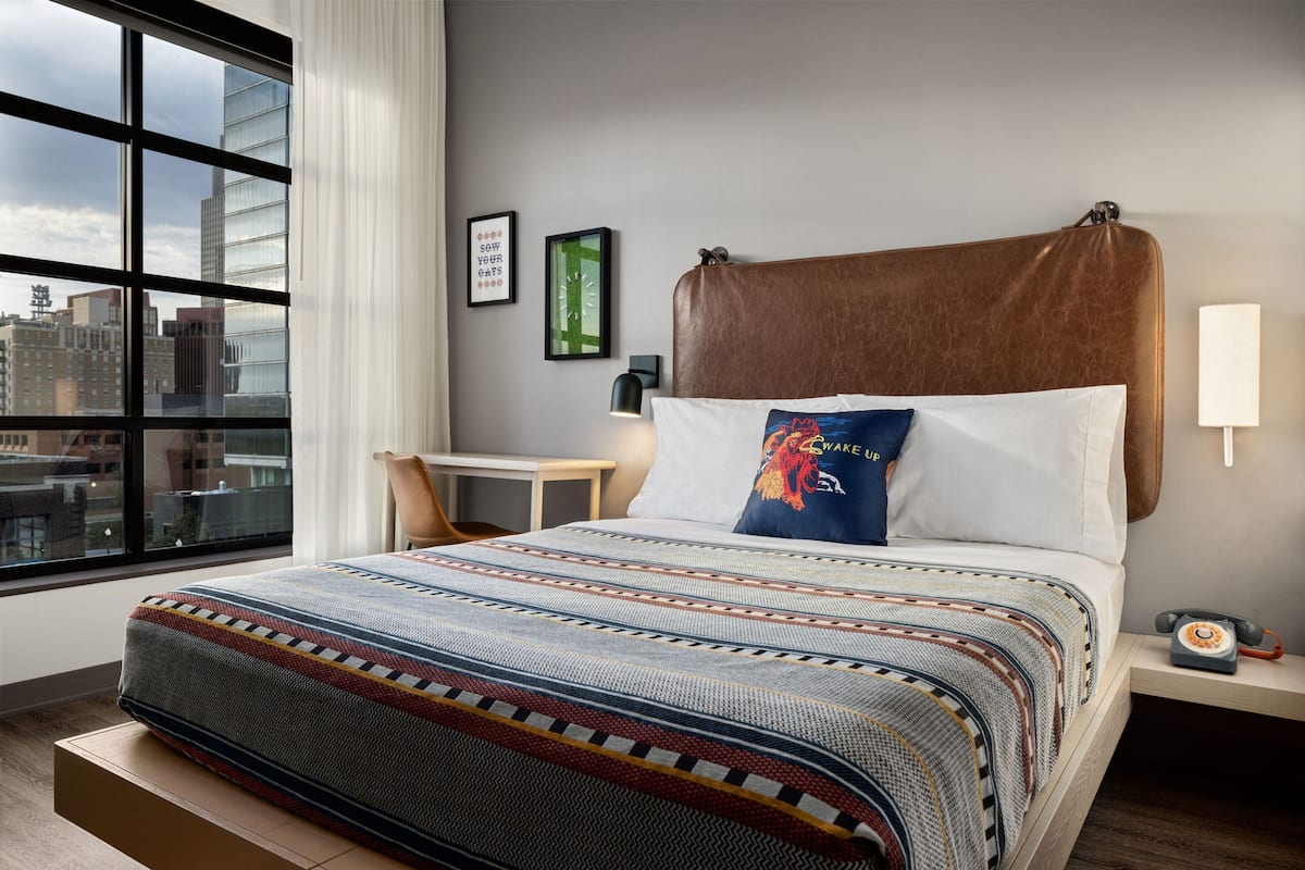 A modern hotel room with a large window, a bed with a patterned bedspread, a headboard, a bedside table with a lamp, and a city view.