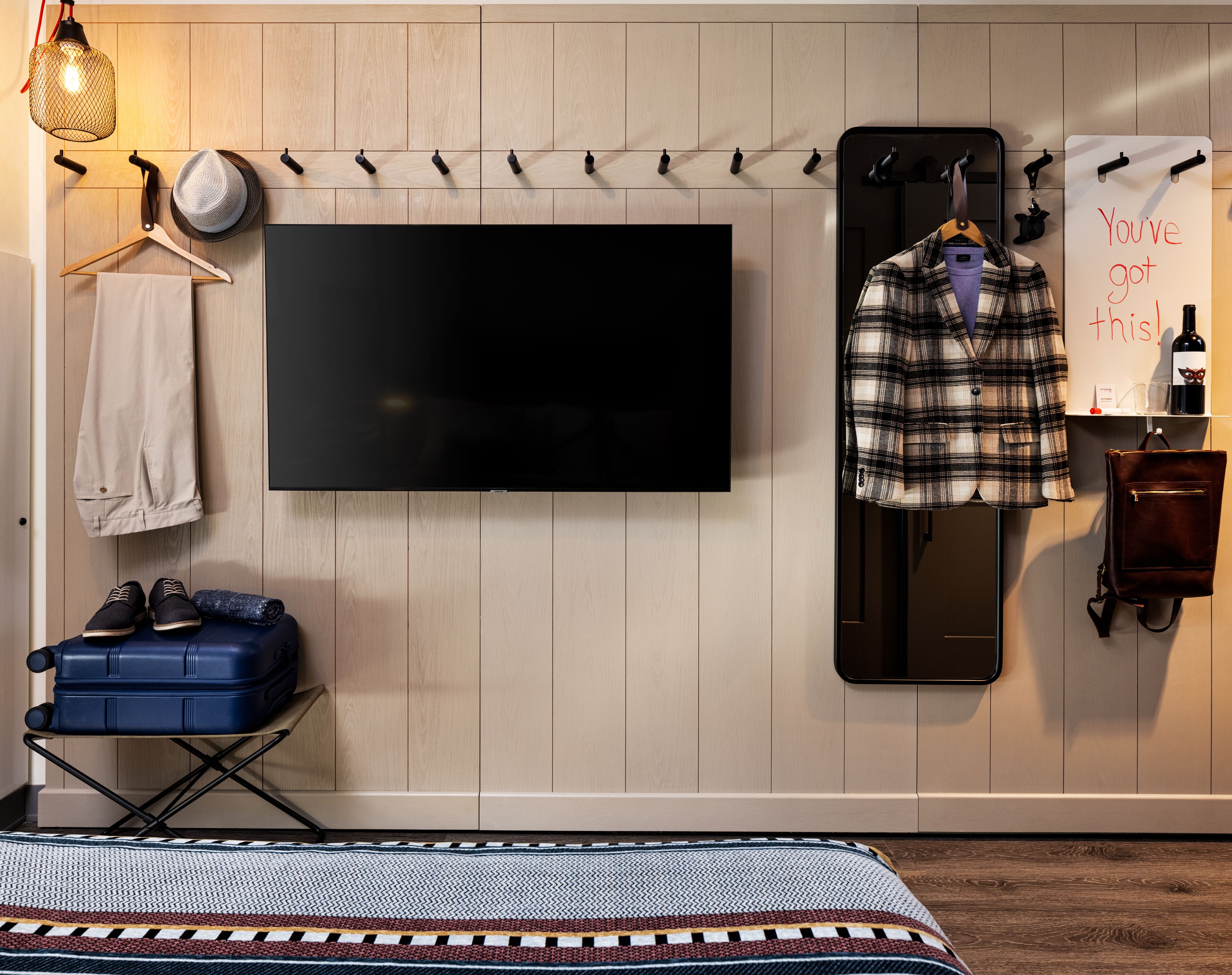 Une chambre d’hôtel équipée d’un téléviseur à écran plat fixé au mur, d’un miroir, d’une valise et d’un porte-manteau mural.
