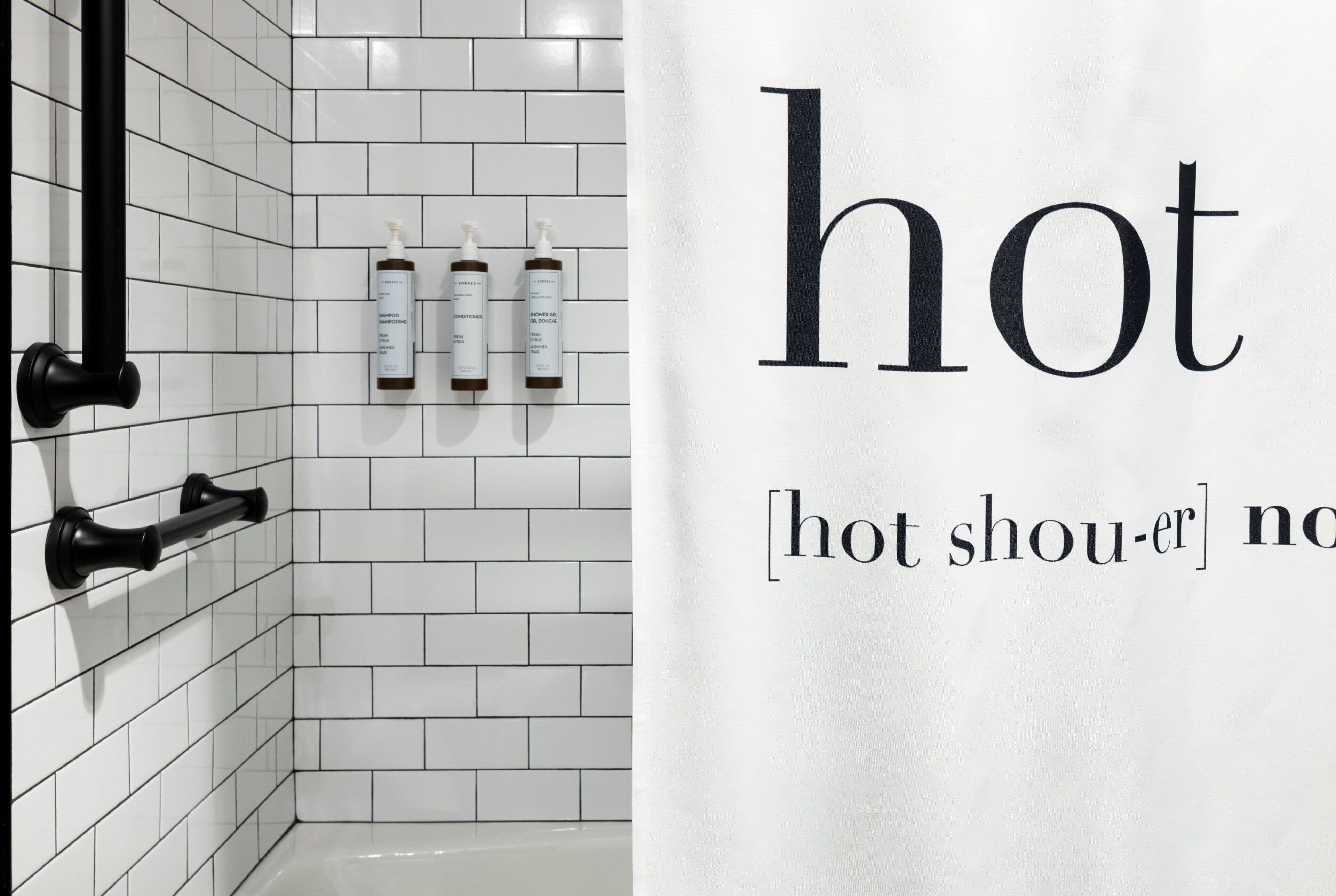 A bathroom with white tiled walls, a black towel rack, and three bottles of shampoo and conditioner.