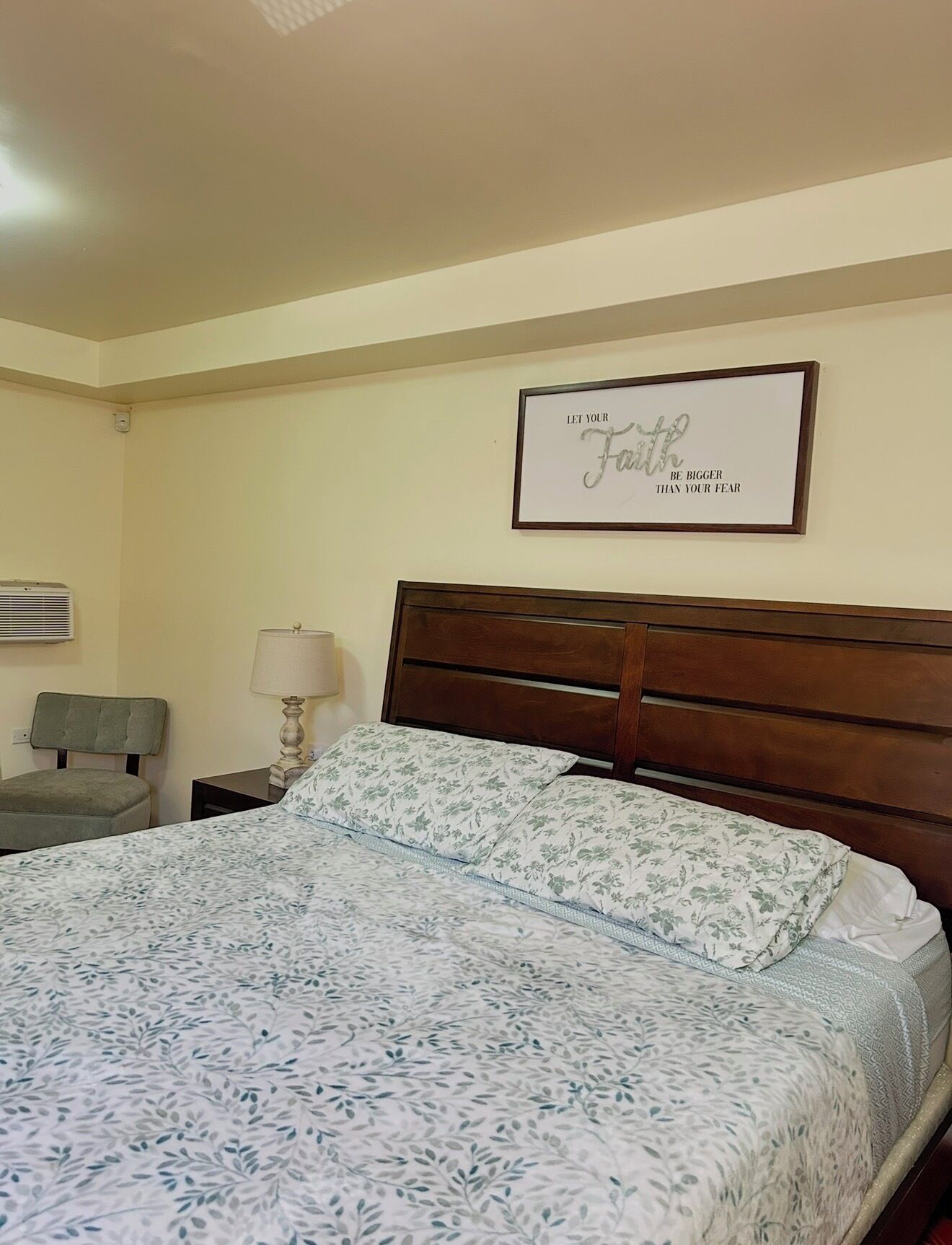 A hotel room with a bed, a nightstand, a lamp, a chair, and a wall-mounted air conditioning unit.