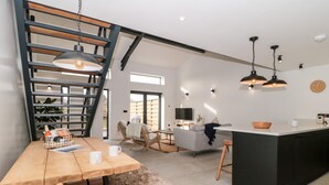 A modern kitchen with a central island, a dining area with a wooden table and chairs, and a staircase with a wooden railing. - Linden Barn (Okehampton)