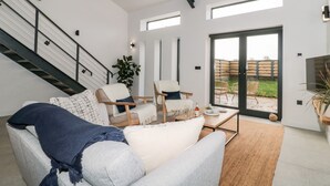 A modern living room with a staircase, a sofa, two armchairs, a coffee table, and a sliding glass door leading to an outdoor area. - Linden Barn (Okehampton)