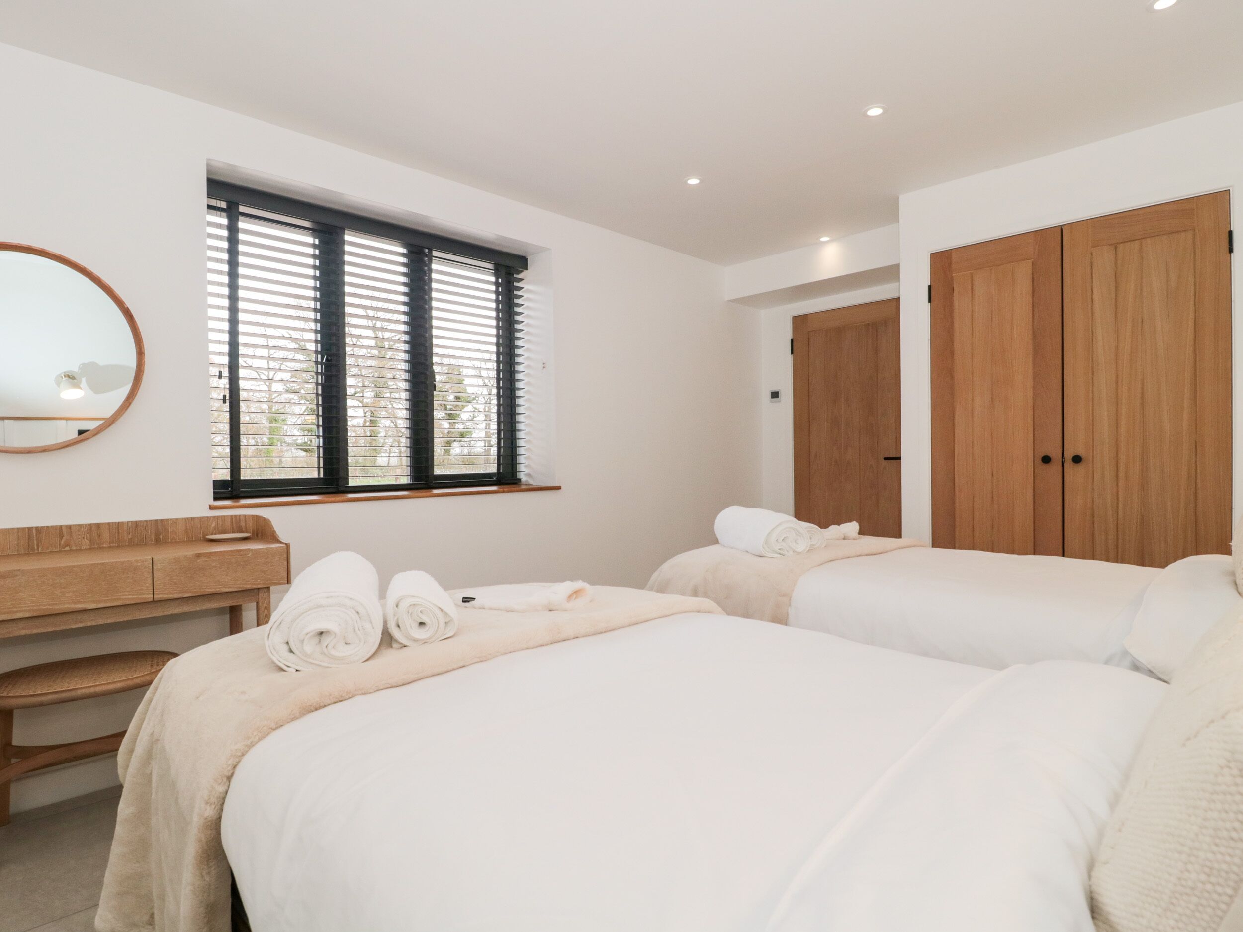 A bedroom with two beds, a wooden bench, a mirror, and a window with blinds.