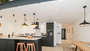 A modern kitchen with a central island, bar stools, pendant lighting, and a dining area with a wooden table and bench. - Linden Barn (Okehampton)