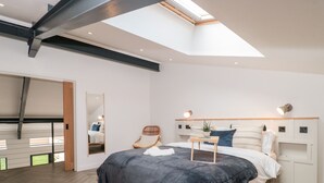 A modern bedroom with a skylight, a bed with a dark blanket, a side table with a plant, and a chair. - Linden Barn (Okehampton)