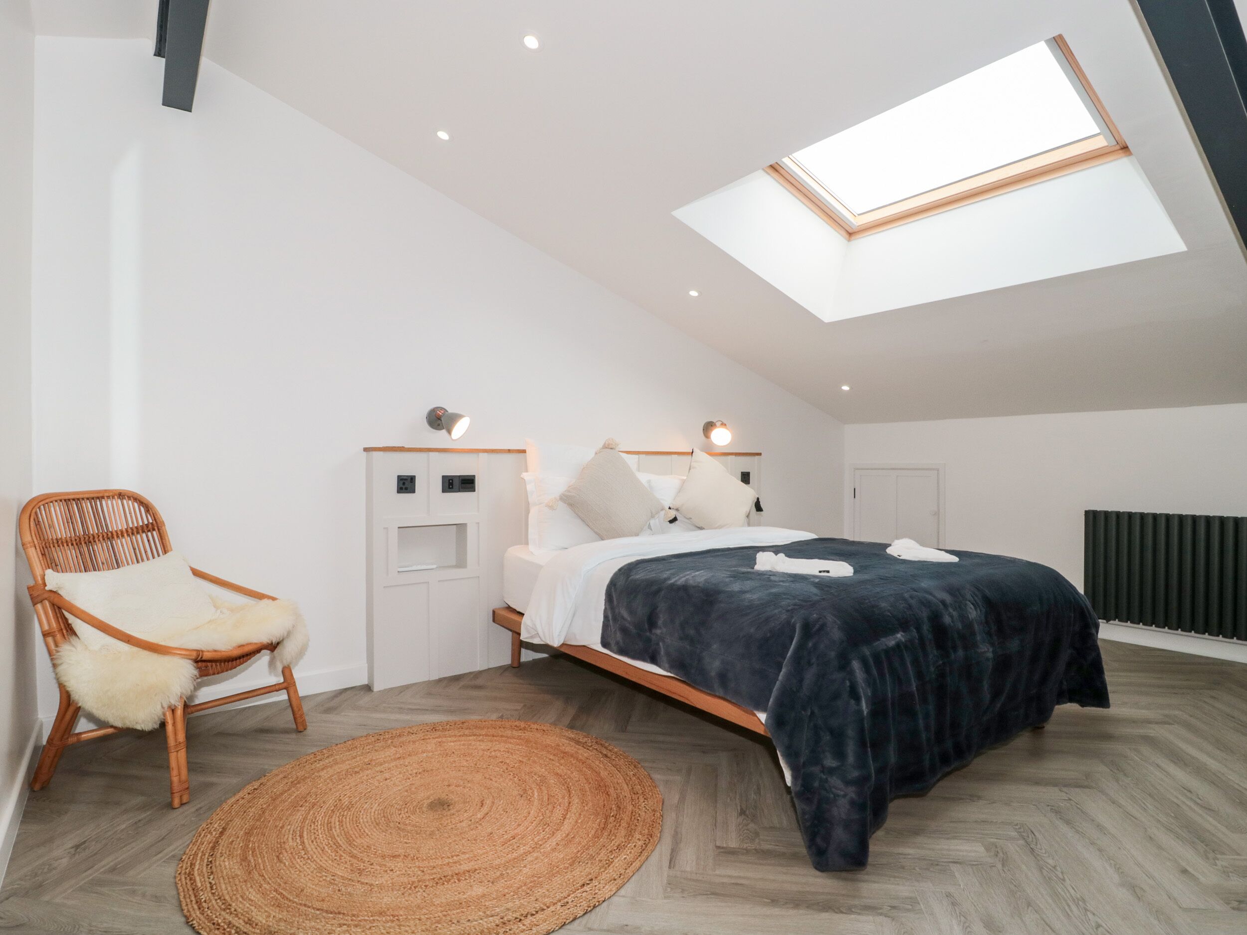 Una camera da letto moderna con un lucernario, un letto con copriletto blu scuro, una sedia in vimini e un tappeto intrecciato.