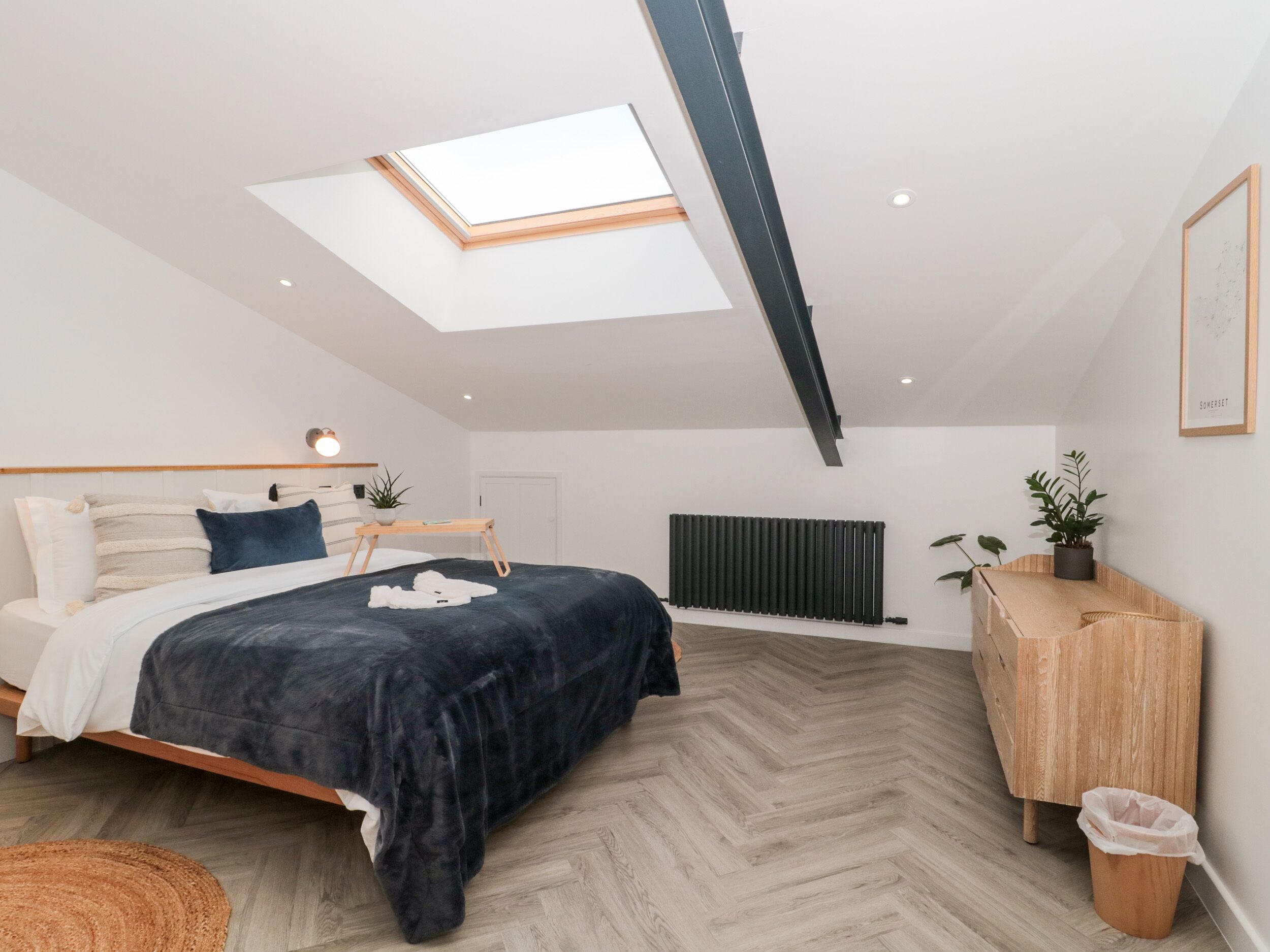 A modern bedroom with a skylight, a bed with a dark cover, a wooden bedside table, a radiator, and a wooden cabinet.