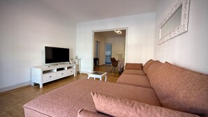 Una sala de estar con un televisor de pantalla plana grande, un mueble blanco para equipo multimedia, un sofá rosa y una pequeña mesa de centro blanca.