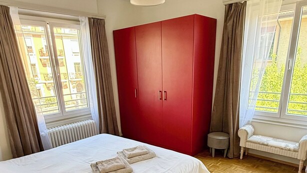 Un dormitorio con cama, un armario rojo, una mesita de noche, un banco junto a la ventana y un radiador.