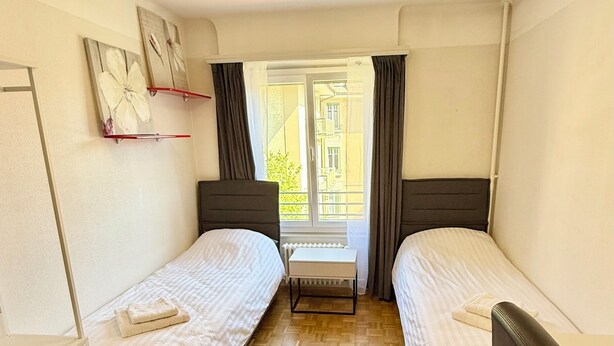 A small, clean room with two single beds, a desk, a chair, and a window with curtains. - CH Apartment Lausanne Gare (Lausanne)